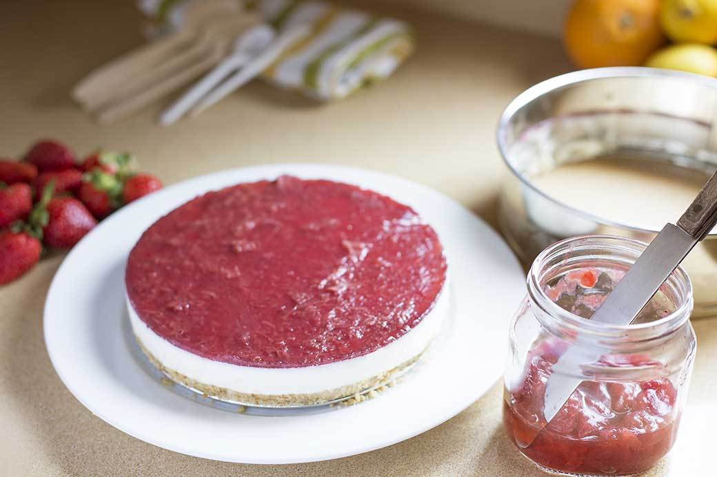A layered desert made in a cake ring sits on a counter next to strawberries and a half-full jar of strawberry jam. The bottom layer of the desert is pastry, the middle layer a white custard, and the top layer is strawberry jam.